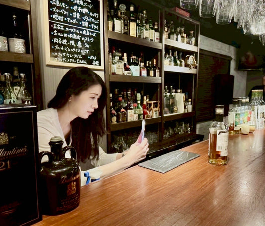 落ち着いた雰囲気のバーのカウンターで、ウイスキーボトルを前にスマートフォンで撮影を行う女性。大人の空間と丁寧な時間の流れが伝わるシーン。