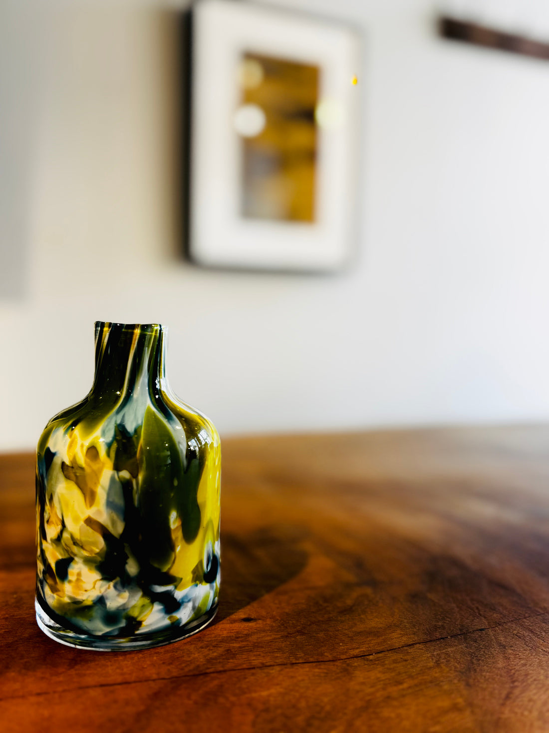 和モダンな空間に置かれた、緑と黄色の模様が美しいガラス花器。木のテーブルと背景の額装アートに調和するインテリア。