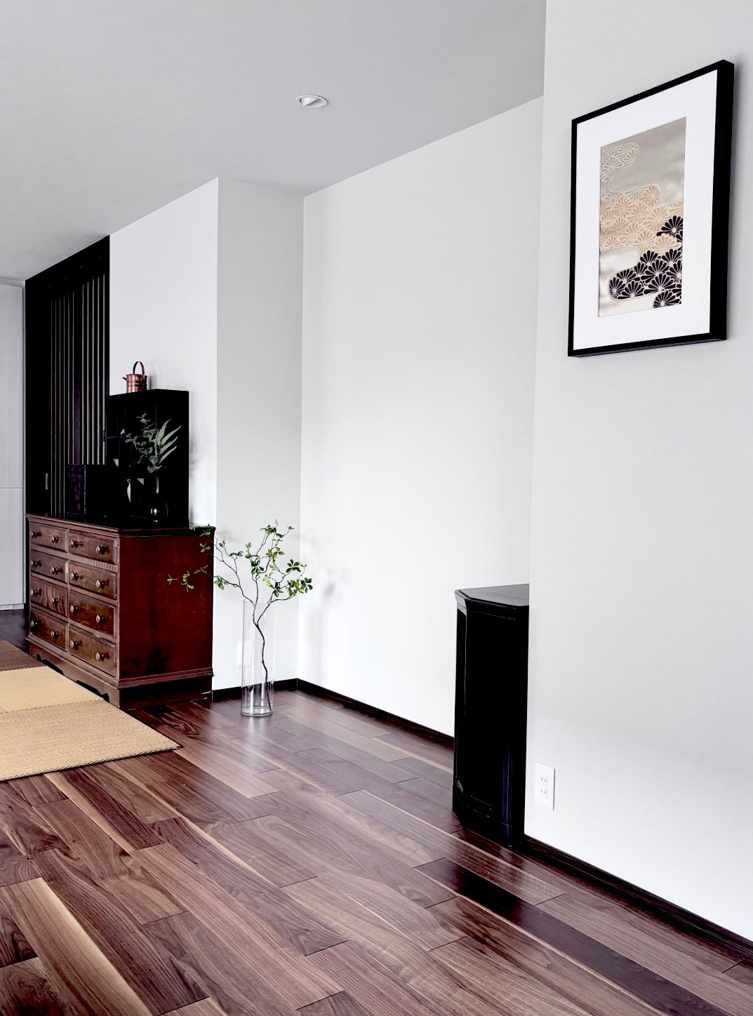 SAKURA JAPANの「帯を切らずに飾る額縁（黒）」を配置した和モダンなリビング・インテリアの全体像 。白い壁面に、上品な金糸や紺色で菊や松などの吉祥紋様が織り込まれたアンティーク帯が、黒いフレームで美しく額装されています 。無垢のウッドフローリング、和の情緒を感じるチェストや格子の建具、ガラス瓶に生けられた緑の枝物ディスプレイと調和し、大切な形見の帯を傷つけずにモダンな壁掛けアートとして愛でる洗練された暮らしのデザイン 。
